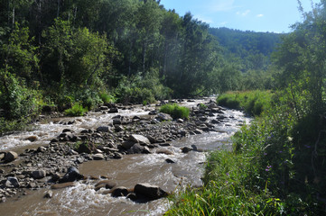 A mountain winding river with rocks and cobblestones amidst green mountains, trees, clouds and blue sky.