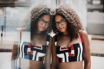 Portrait of an appealing female hipster in eyeglasses with bulky curly hair leaning against glass...