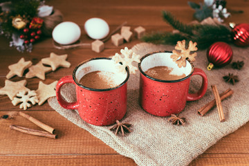Traditional Christmas drink eggnog with cinnamon, anise stars and grated nutmeg in red mugs on the wooden table. Gingerbreads, eggs, and fir-tree decoration