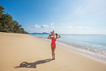 Happy young woman with long hair in red swimsuit wearing funny christmas deer horns holding her hair like two ponytails standing on the beach by the sea with blue water on Phuket island,Thailand