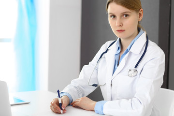 Doctor woman filling up prescription or medical record form while  sitting near window in clinic or hospital. Medicine and healthcare concept. Physician at work