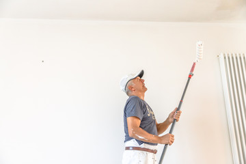 Whitewasher painter man with paint stick roller in hand inside an empty renovation room for painting in white. White wall copy space.