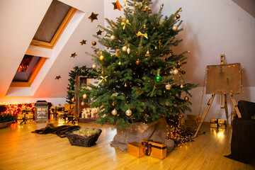 Christmas decorations in the studio, big natural fir tree with golden balls, bows and snowflakes, wooden fireplace and candles on it and brown cozy armchair