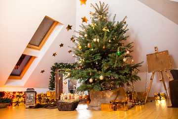 Christmas decorations in the studio, big natural fir tree with golden balls, bows and snowflakes, wooden fireplace and candles on it and brown cozy armchair