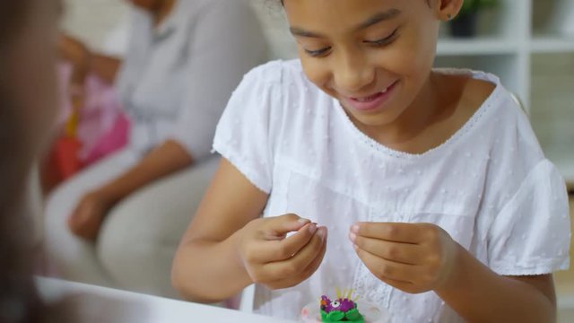 Tilt down shot of cute black girl of primary school age making hedgehog craft with modeling clay and pasta, and smiling