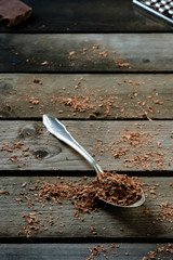 Nahaufnahme, geraspelte Schokolade auf Silberlöffel, Reibe und Schokostück im Hintergrund, verwitterter Holzuntergrund