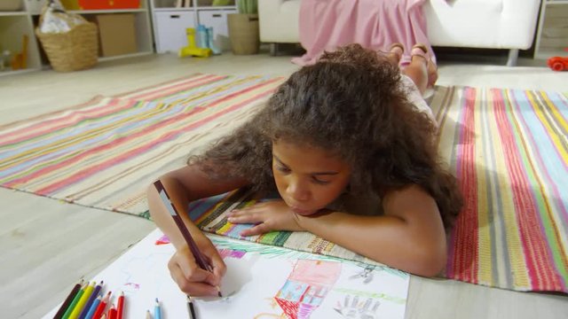 Bored little black girl lying on rug at home and drawing picture with pencils of different colors