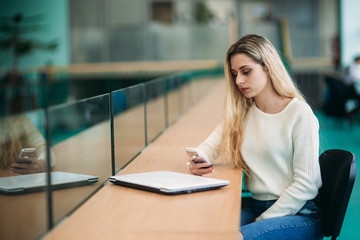 Blonde girl use phone in library. sudent sist on stool by the window