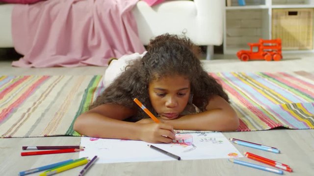 Front view of concentrated black girl of elementary school age lying on rug and drawing picture with pencils of different colors