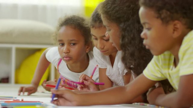 Medium shot of group of little black children lying on floor and drawing pictures with colored pencils, tilt up