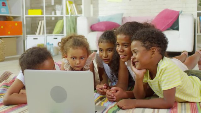 Group of interested black children of elementary school age lying on rug at home, watching something together on laptop computer and smiling