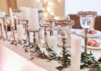 Wedding table serivce. Candles on silver vases and glasses with water stand among white flowers on wedding dinner table