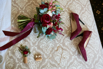 Wedding decor. Classy bouquet of roses, dark pink shoes and golden accessories for a bride lie on the table