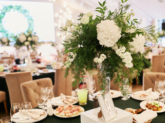 Wedding reception decor. Round dinner tables covered with pink and green clothes and served with glasses, crockery and white flowers stand in the hall