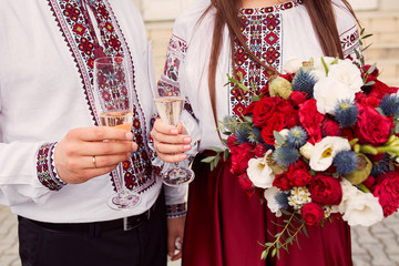 Fototapeta na wymiar Bride and groom in ethnic Ukrainian clothes clang glasses with champagne