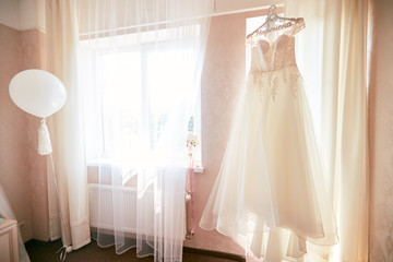 Rich white wedding dress hangs before a window in the hotel room