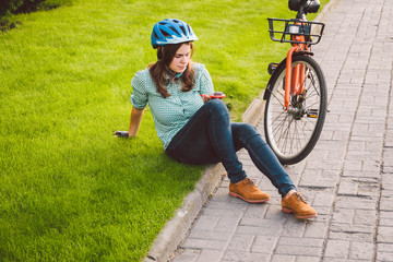Man and city rolling bicycle, environmentally friendly transport. Beautiful young caucasian woman worker sitting resting on the grass uses a red mobile phone near an orange bicycle with a coryne.