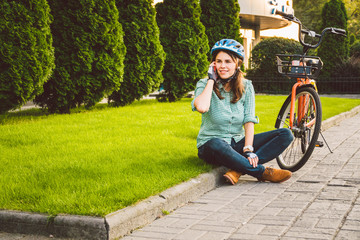 Man and city rolling bicycle, environmentally friendly transport. Beautiful young caucasian woman worker sitting resting on the grass uses a red mobile phone near an orange bicycle with a coryne.