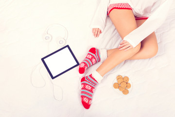 Obraz na płótnie Canvas Young woman in Christmas cozy socks, sitting in bed, with tablet, headphones and cookies next to her
