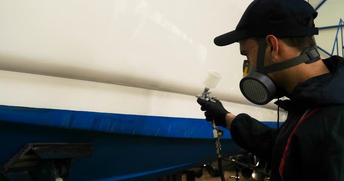 Professional master man in a robe and puller with spray gun puts a layer of paint or ceramics on the yacht. Concept of: Dangerous work, Chemistry, Paints, Garage, Transport Service.