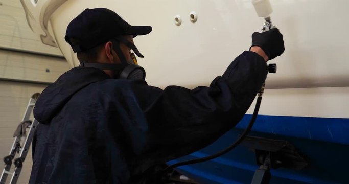 Professional master man in a robe and puller with spray gun puts a layer of paint or ceramics on the yacht. Concept of: Dangerous work, Chemistry, Paints, Garage, Transport Service.