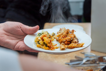 Closeup of a Hand Holding a Plate of Steaming Hot Pasta with Vegetables and Cheese