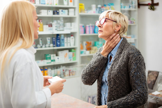 Tired, ill mature woman holding her throat and explaining problems to a beautiful young female pharmacist. Thyroid gland problems. Medicine, healthcare and pharmaceutics concept