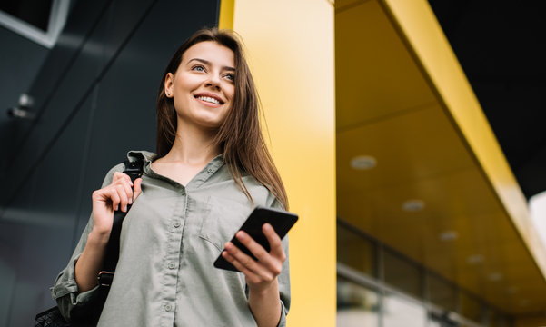 Confident happy businesswoman with beautiful smile and face using modern mobile phone, chatting, thinking and planning creative project standing near business centre. Successful business. Career.