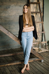 Portrait of beautiful smiling brunette girl posing against of wooden textured wall barefoot near ladder looking away with crossed arms in blue jeans with jacket and vest on shoulders. Fashion pose.