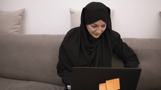 Happy, smiling muslim woman in black clothes sitting on a grey couch at home and working with her laptop on her knees, typing, freelancing at home, talking to camera. Loft grey room interior
