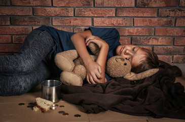 Poor homeless boy sleeping with his toy on floor near brick wall
