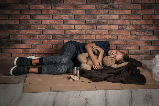 Poor homeless boy sleeping with his toy on floor near brick wall