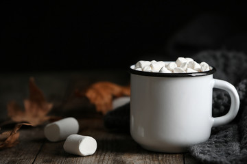 Mug of hot cozy drink with marshmallows, sweater and autumn leaves on table. Space for text