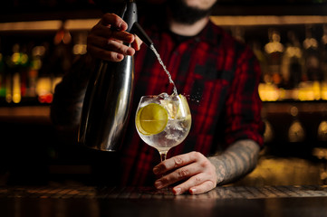 Bartender adding tonic water to the to the cocktail glass full of ice cubes with lime slices