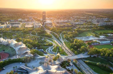 Modernes europäisches Stadtbild aus der Luft mit Gewerbe- und Sportanlagen, Parks, mehrspuriger Autobahnkreuzung, Hochhäusern in Außenbezirken, die in der Abenddämmerung von warmem Sonnenlicht beleuchtet werden München Bayern Deutschland © nogreenabove2k