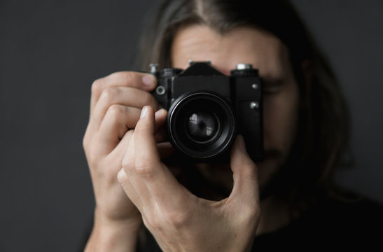 Handsome young bearded man with a long hair and in a black shirt holding vintage old-fashioned film camera on a black background and looking in camera viewfinder.