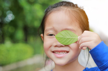 Adorable little Asian child girl holding a green leaf closing left eye in green garden background.