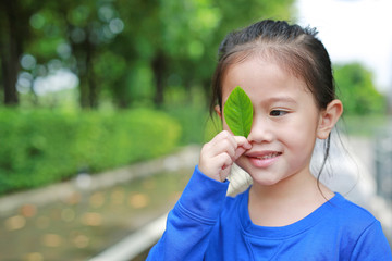 Adorable little Asian child girl holding a green leaf closing right eye in green garden background.