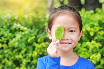 Adorable little Asian child girl holding a green leaf closing right eye in green garden background.