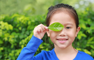 Close up little Asian child girl holding a green leaf closing right eye in green garden background.