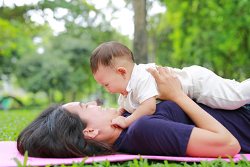 Happy Asian mom embrace her son lying in the green garden. Funny mother and infant baby boy playing in summer park.