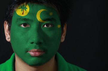 Portrait of a man with the flag of the Cocos (Keeling) Islands painted on his face on black background, a palm tree on a gold disc, crescent and southern cross on green.