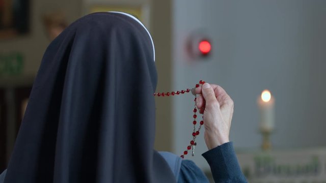Religious sister praying on rosary beads with red tabernacle light on the wall in the background and a candle on the altar.