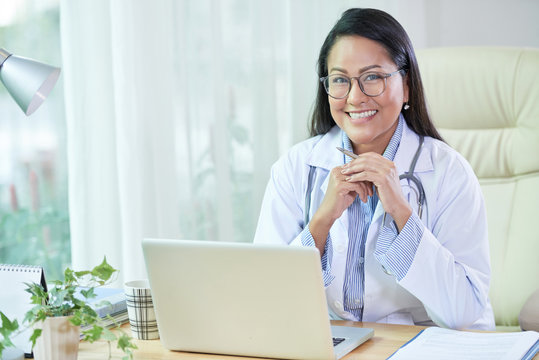 Beautiful Adult Thai Woman In Doctor Coat And Glasses Sitting At Table With Laptop Smiling At Camera