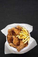 Tasty fastfood: fried chicken drumsticks, spicy wings, French fries and chicken fingers in paper box over black background, top view. Flat lay, overhead, from above. Copy space.