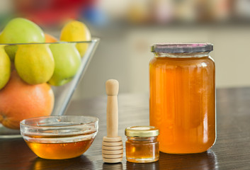 Big and small glass jar of honey with wooden sipper spoon and bowl of fruits on the background.
