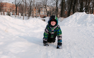 Fototapeta na wymiar Boy plays in the snow. Crawls along a snowy path among snowdrifts in the winter city park.