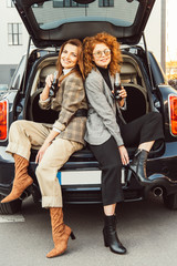 cheerful female friends in jackets posing with soda bottles and sitting back to back in car trunk at urban street