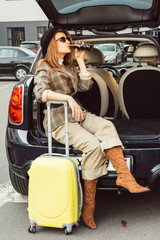 stylish female tourist in black hat drinking soda while sitting in car trunk near wheeled bag at...