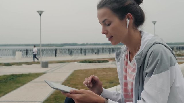 Young woman using tablet outdoor laying on grass, smiling. student after university in campus girl in airpods headphones earpods happy homework